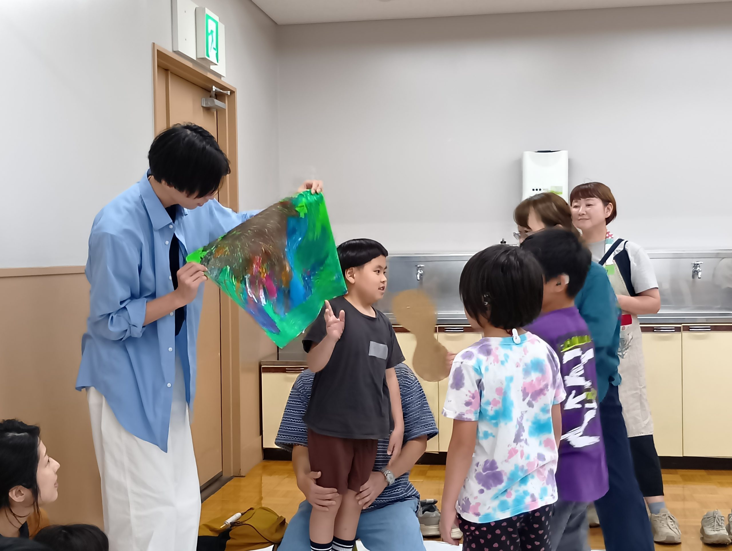 画用紙一面にたくさんの色を塗った男の子が、自分の作品の前で紹介している様子と、それを間近で見る他の子どもたち
