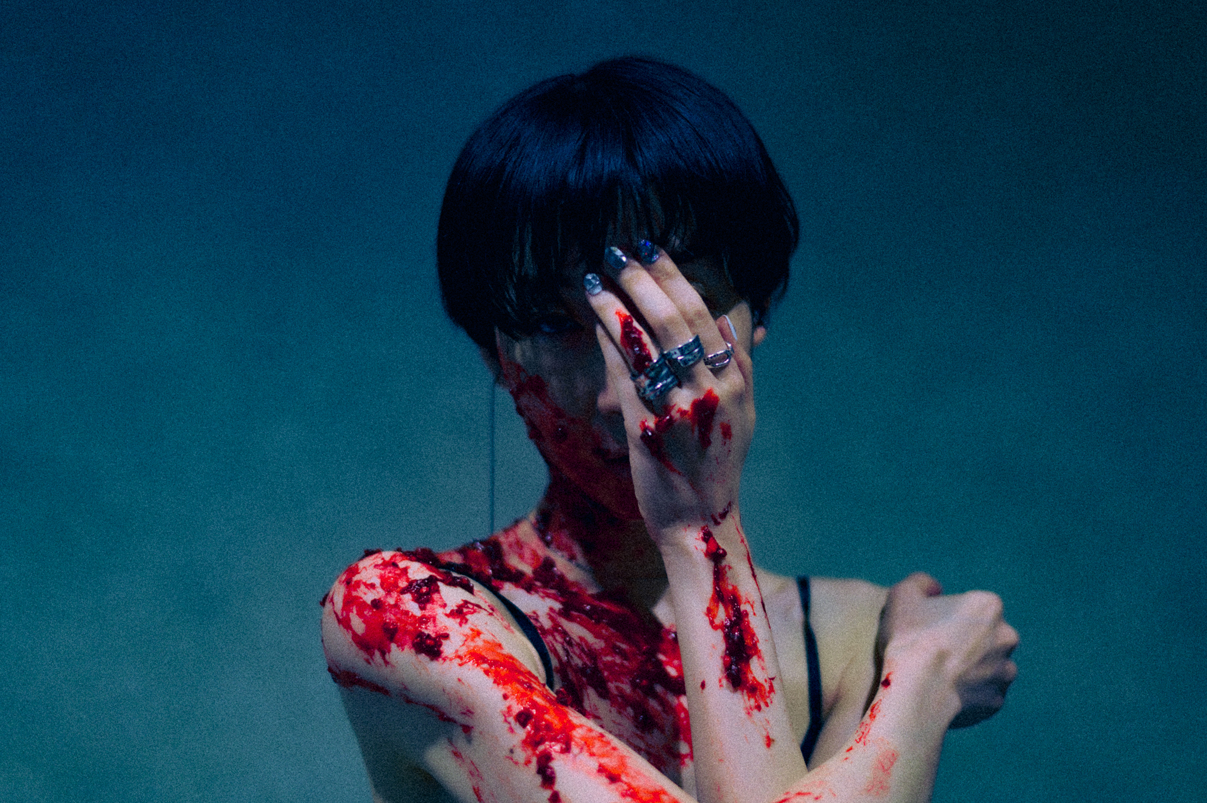 ショートヘアの作家の写真。左手で顔を多い、右手で左肩を支えるポーズをしている。黒いキャミソール姿で、血をおもわせる赤い液体が肌の上を流れている