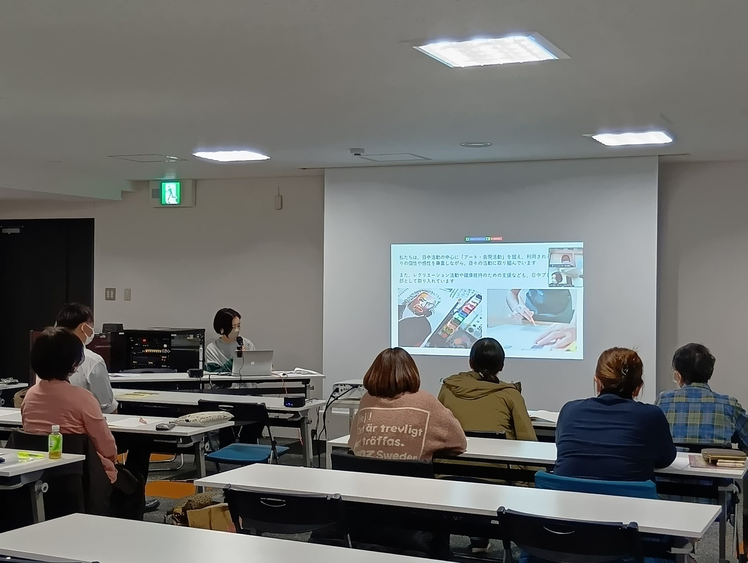 会場の様子の画像。参加者と講師が写っている。研修室の前方のプロジェクターにスライドが投影されている。