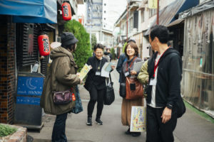 背景に赤い提灯や古い風合いの居酒屋が並び、4人が仲よさそうに会話しながら歩いている様子。