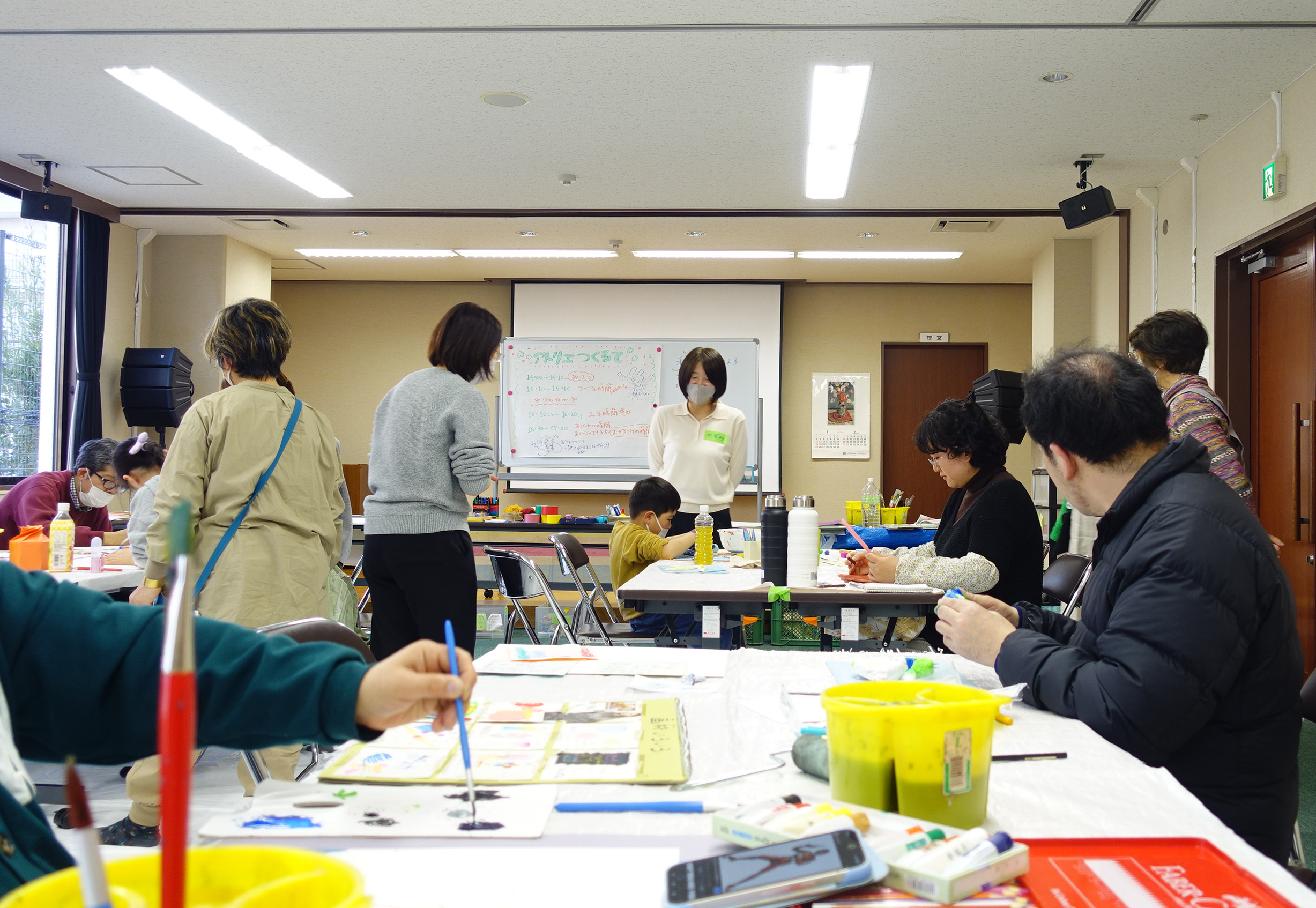 アトリエの風景。テーブルに絵の具で絵を描いている大人、粘土をで立体をつくっているこども、鏡を見て自画像を描いている人などがいる。