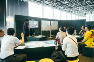黒板の風合いをした机やイスが並んでおり、会場の前で人がモニターを使いながら説明をしている。